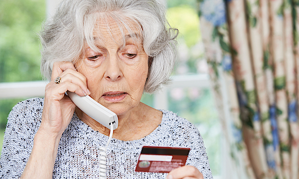 Eine ältere Frau mit grauen Haaren telefoniert. Sie macht ein besorgtes Gesicht. In der Hand hält sie ihre Bankkarte. Sie gibt dem Anrufer ihre Bankdaten durch.